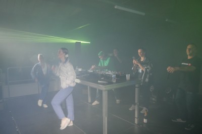 Miniature Equinoxe #39 Un DJ mixe sur une scène éclairée, avec un écran lumineux affichant des motifs rouges en arrière-plan.