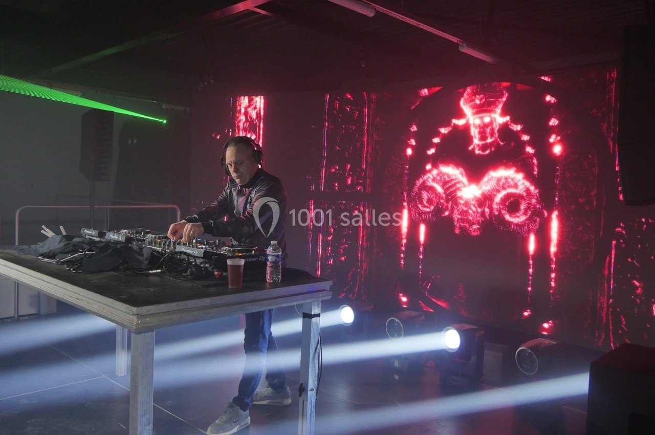 Un DJ mixe sur une scène éclairée, avec un écran lumineux affichant des motifs rouges en arrière-plan.