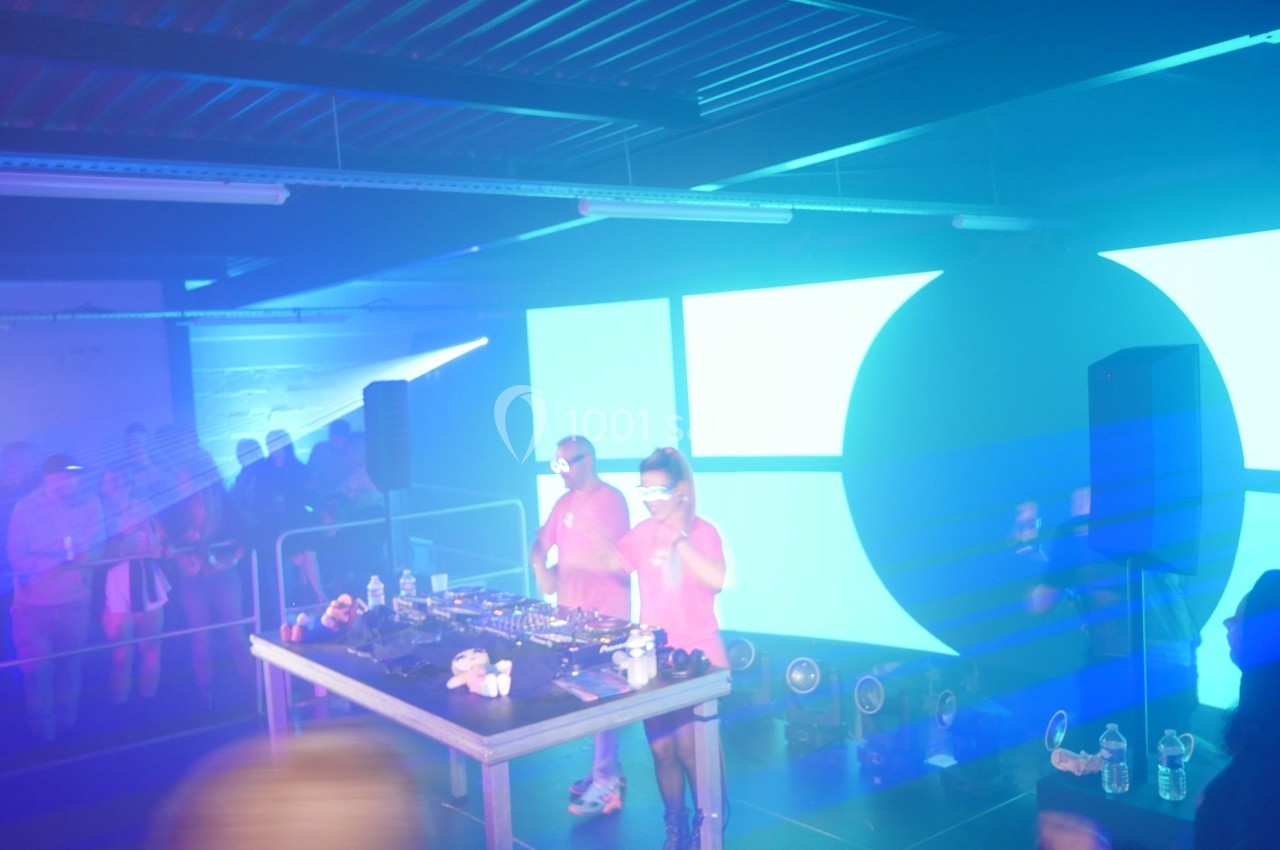 Deux DJ en chemises roses mixent sur une scène éclairée par des lumières bleues et un écran lumineux en arrière-plan.