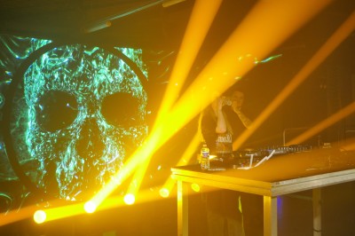 Miniature Equinoxe #42 Un DJ mixe sur scène avec des jeux de lumière jaunes et un écran affichant une image de crâne en arrière-plan.