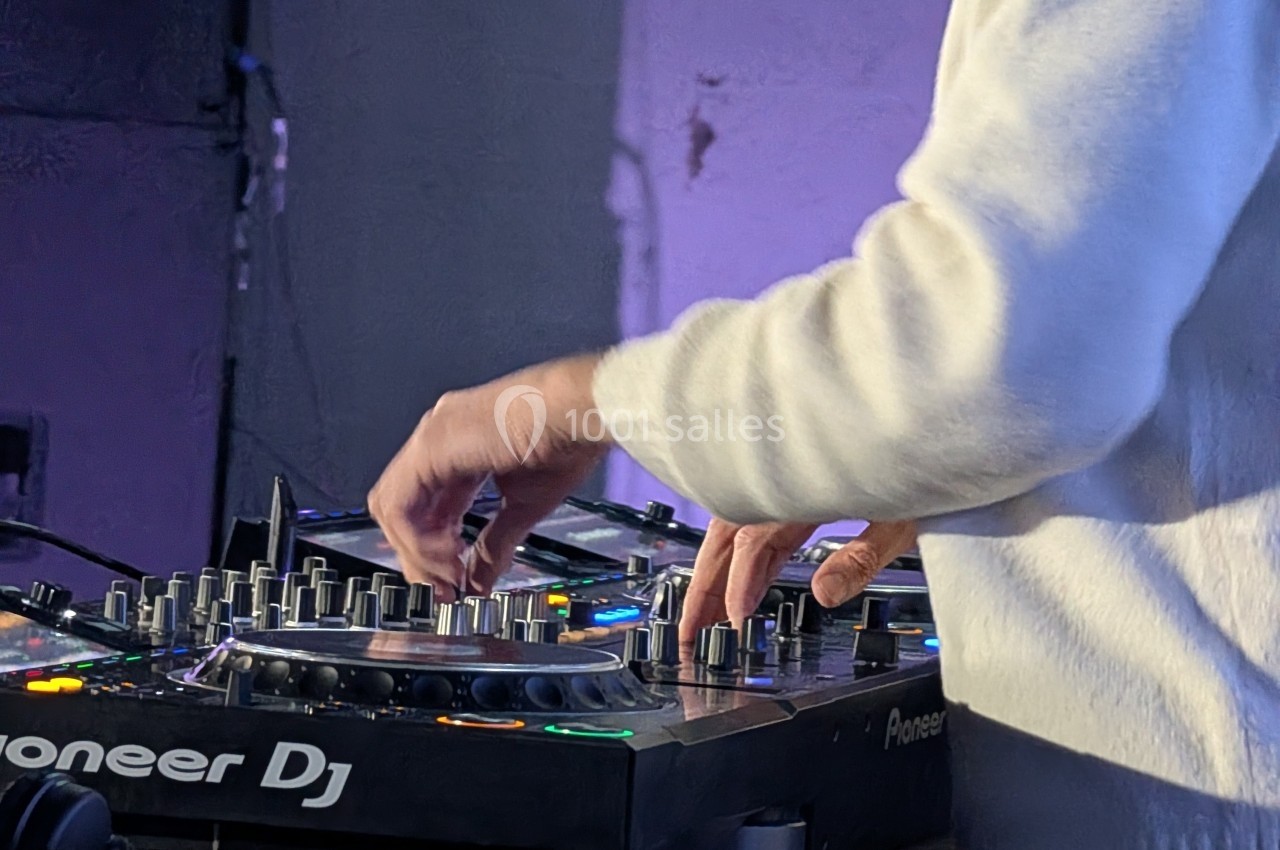 Un DJ en pull blanc manipule une table de mixage avec des lumières colorées dans un environnement sombre.