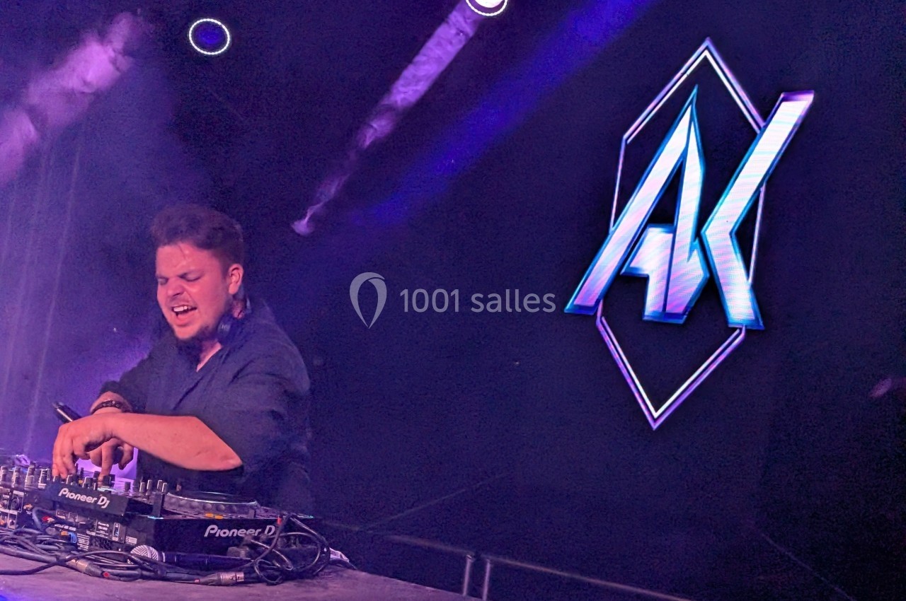 Un DJ mixe sur scène, entouré de lumières violettes, avec un logo lumineux ’AK’ en arrière-plan.