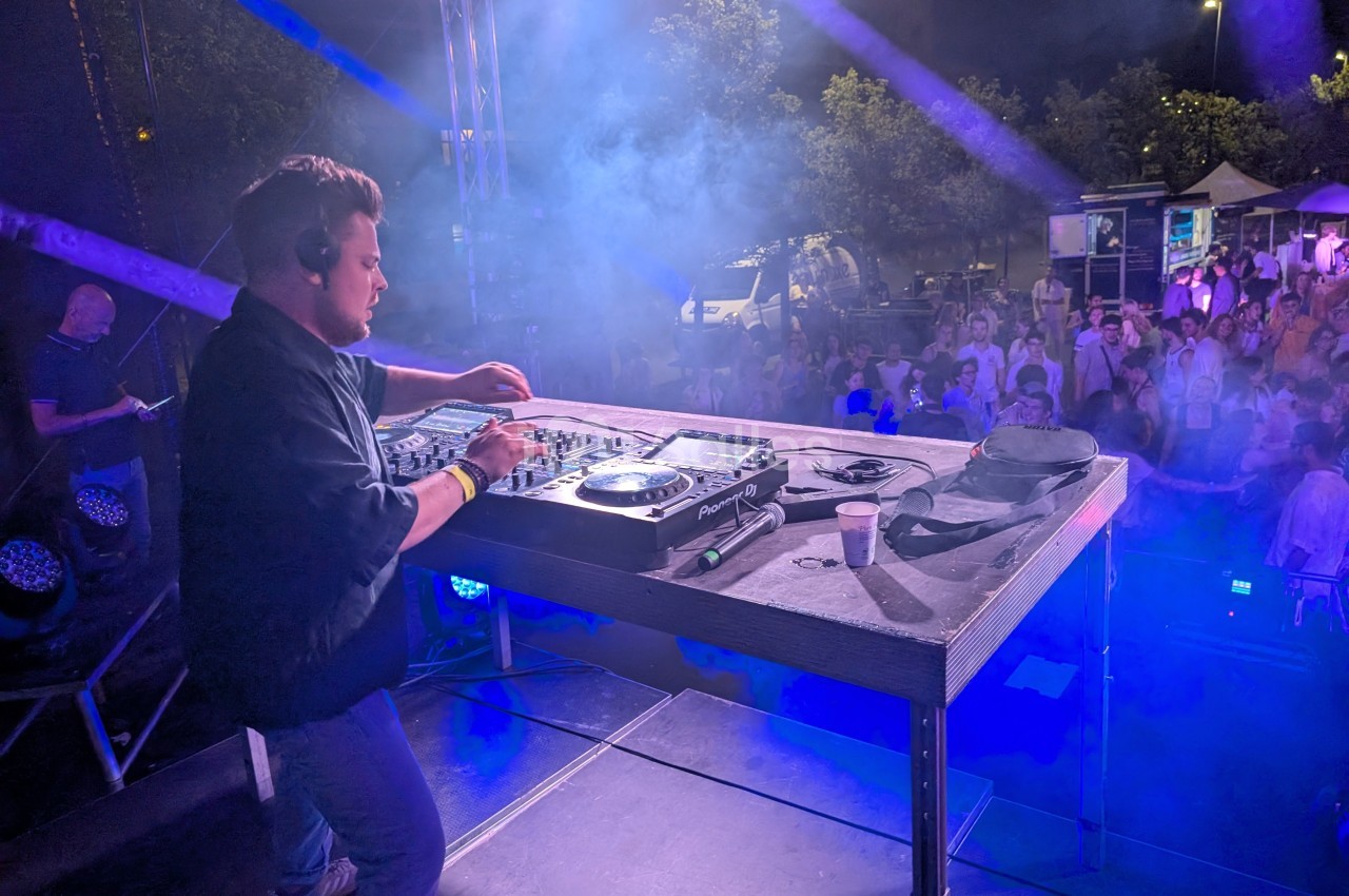 Un DJ mixe sur une scène en plein air, face à une foule sous des lumières colorées, de nuit.