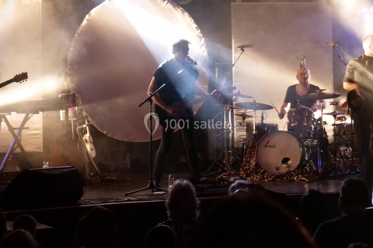 Un groupe joue sur scène dans une ambiance lumineuse tamisée, avec un batteur, un guitariste et des spectateurs en ombre.