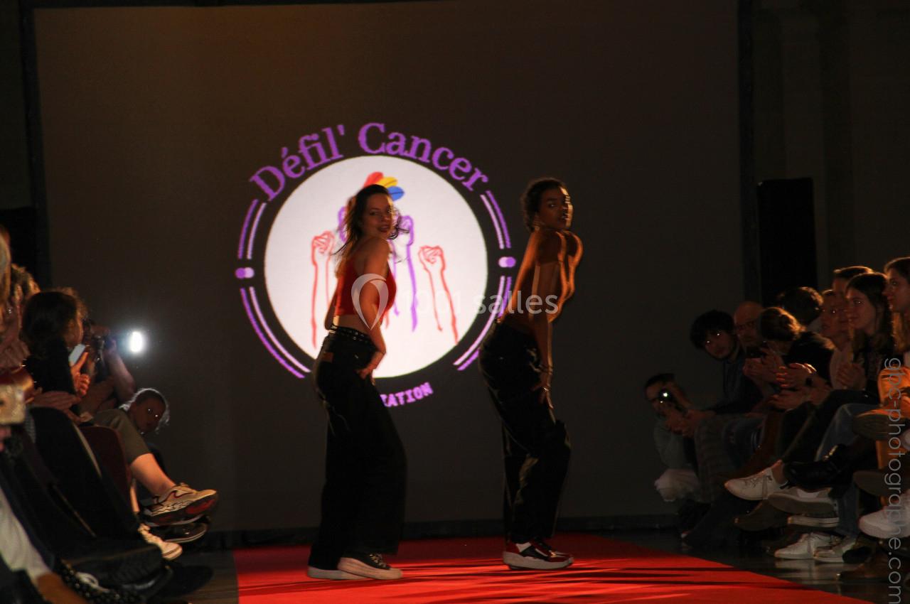 Deux personnes dansent sur un podium éclairé, devant un écran affichant ’Défil' Cancer’, entourées de spectateurs.
