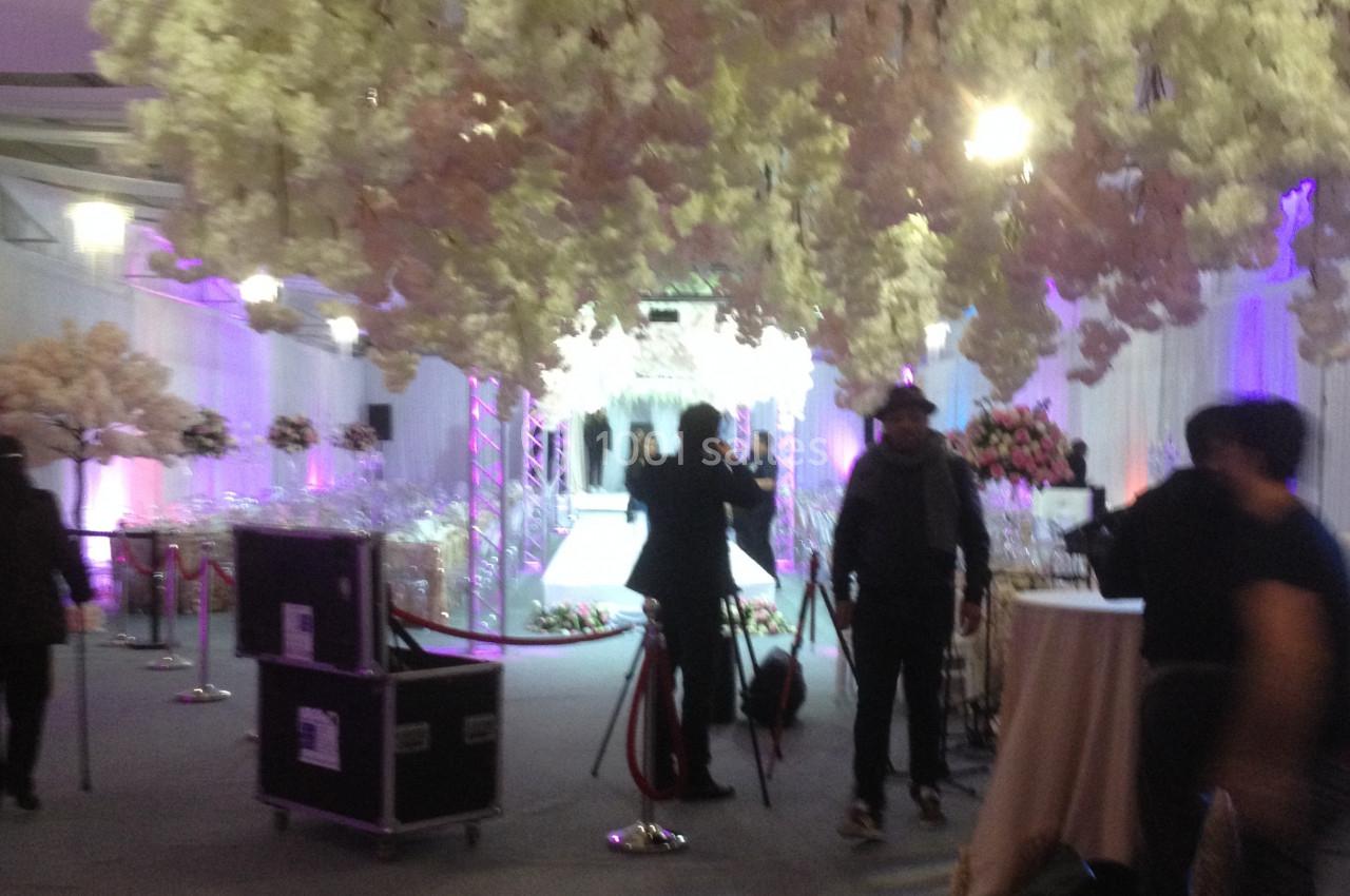 Salle décorée avec des fleurs suspendues, éclairage tamisé et personnes installant du matériel audiovisuel.