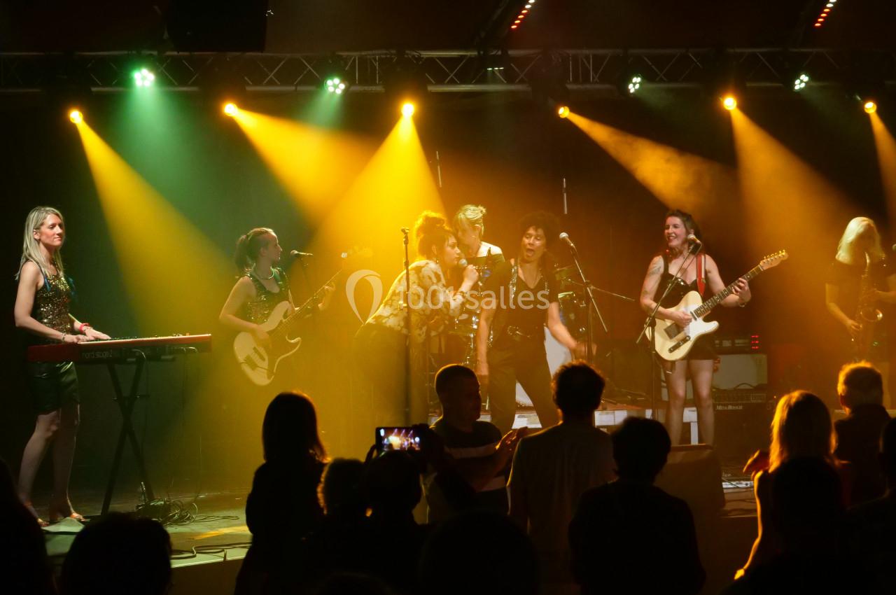 Groupe de musiciens et chanteurs sur scène, éclairés par des projecteurs colorés, devant un public debout.