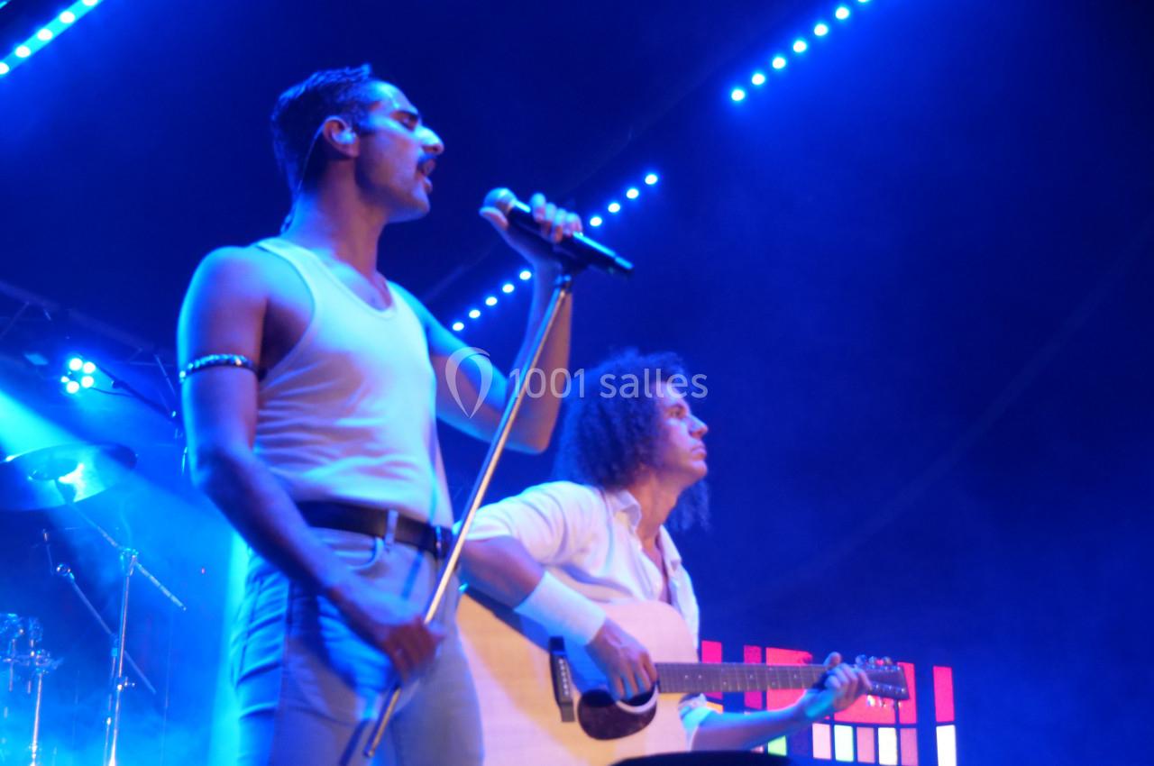 Deux musiciens sur scène, l'un chantant avec un micro, l'autre jouant de la guitare sous des lumières bleues.