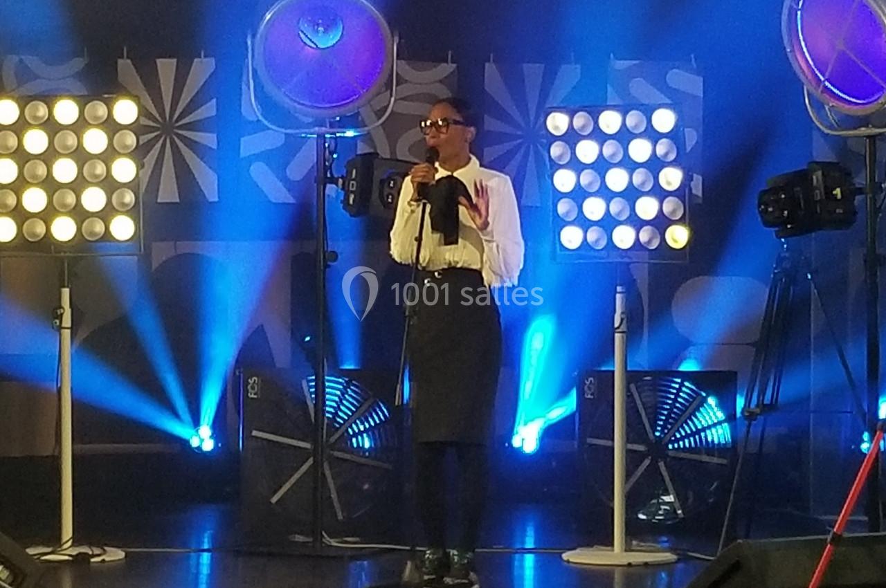 Une personne chante sur une scène éclairée par des projecteurs, entourée d'équipements audiovisuels.