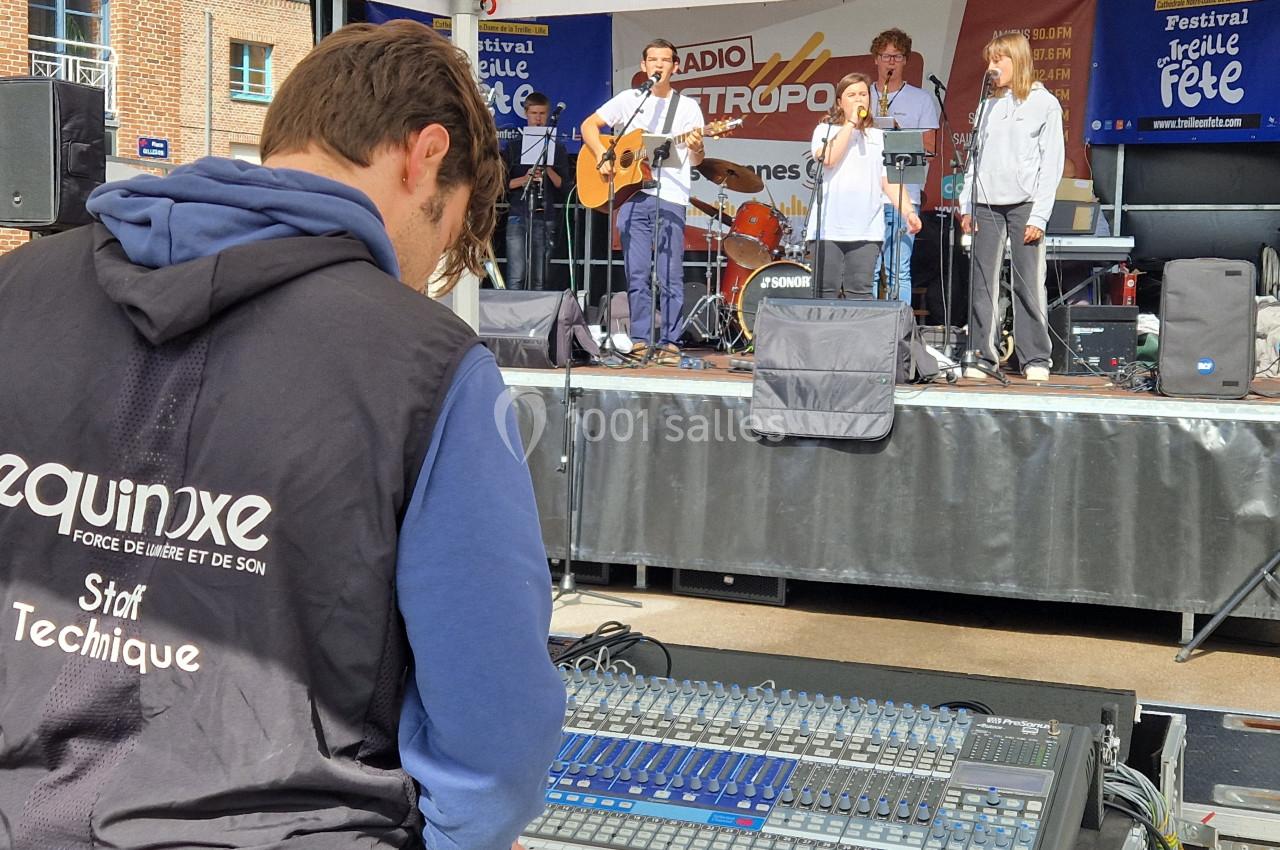 Un technicien ajuste une console de mixage lors d'un concert en plein air avec un groupe jouant sur scène.