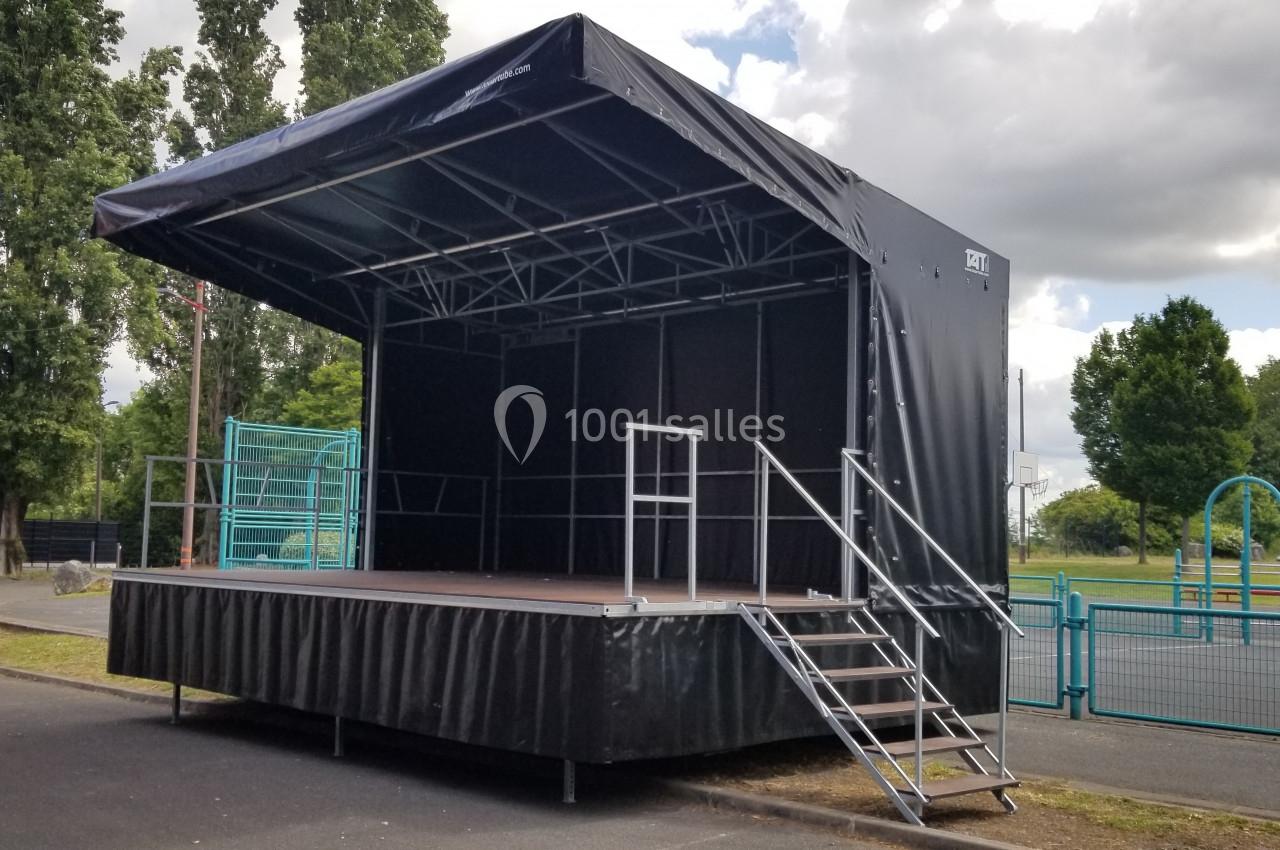 Scène mobile extérieure avec structure métallique et bâche noire, installée dans un espace ouvert près d'une aire de jeux.