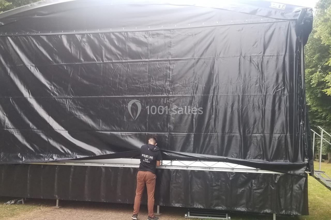 Un homme ajuste la bâche noire d'une scène extérieure montée dans un espace arboré.