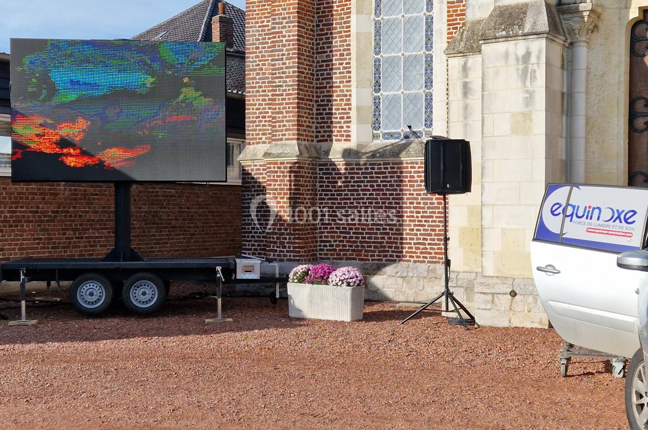 Écran géant monté sur une remorque devant un bâtiment en briques, avec une voiture stationnée à droite.