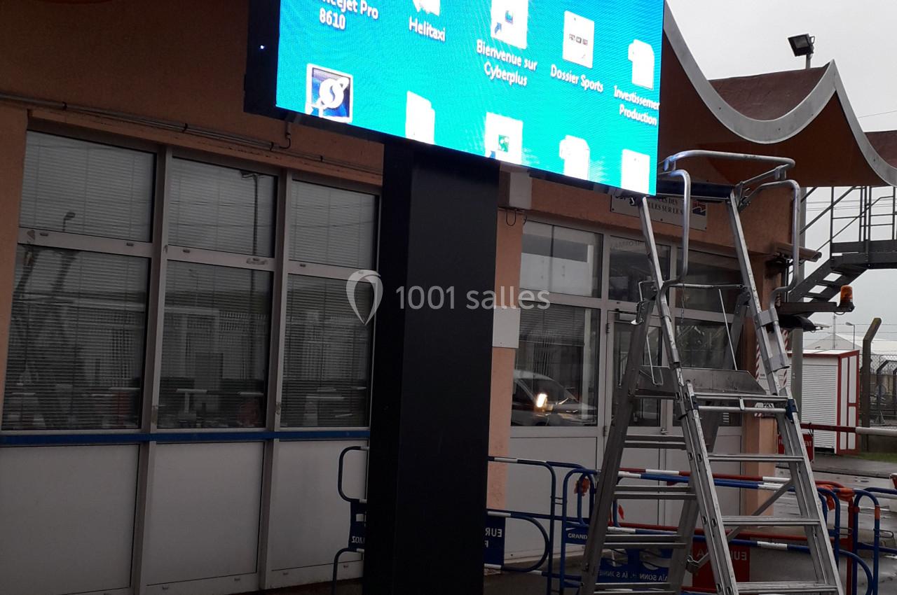 Écran numérique monté sur un poteau devant un bâtiment, affichant une interface d'ordinateur avec des icônes de dossiers.