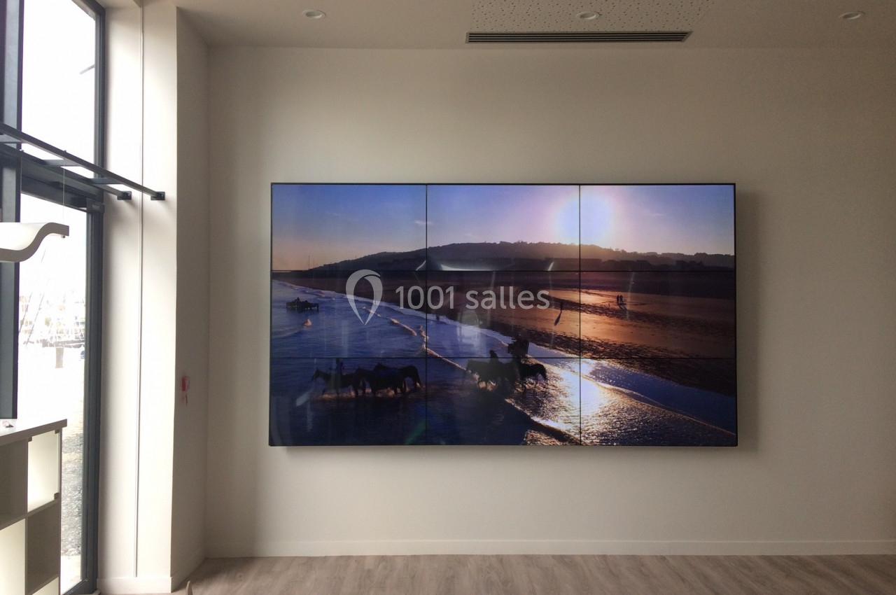 Écran mural affichant une plage au coucher de soleil, dans une pièce lumineuse avec sol en bois et grandes fenêtres.