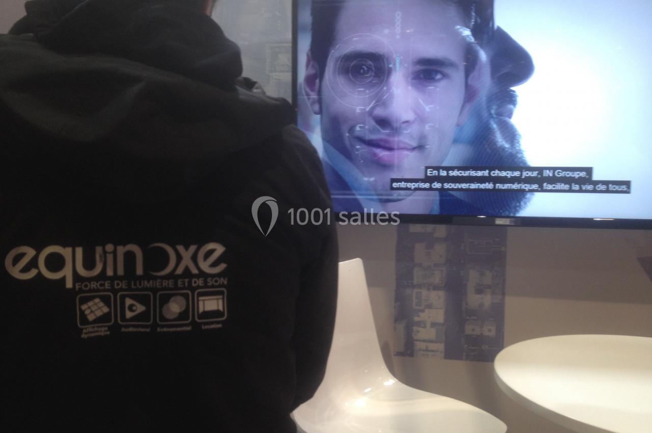 Une personne de dos portant un vêtement marqué ’equinoxe’ regarde un écran affichant un visage et des sous-titres.