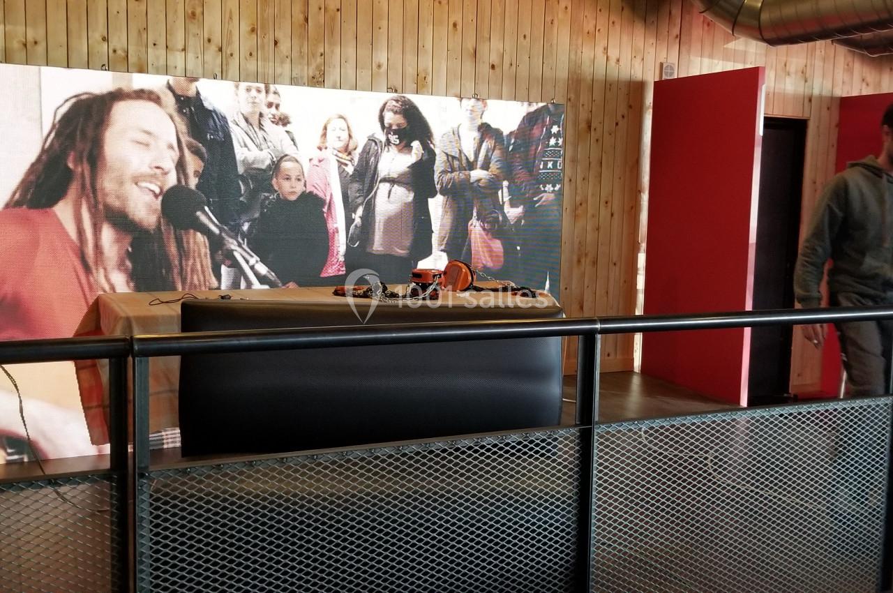 Un écran affiche un chanteur en concert, installé derrière une table avec des équipements, dans une salle en bois.