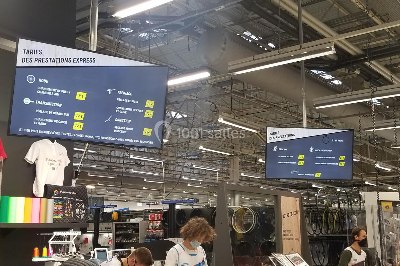 Écrans affichant les tarifs des prestations dans un atelier de réparation, avec employés et clients en arrière-plan.