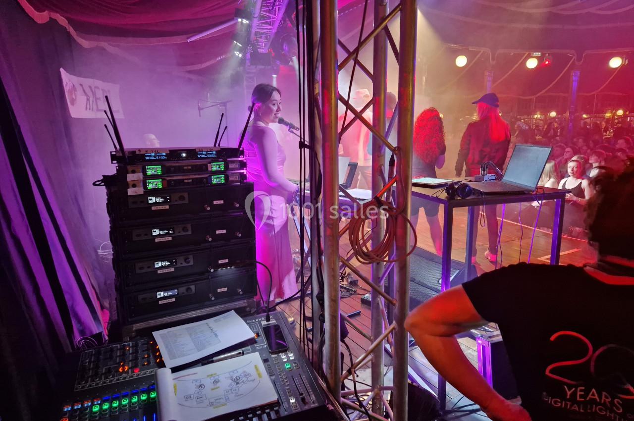 Vue des coulisses d'une scène avec des équipements audio, des artistes sur scène et un public éclairé en arrière-plan.