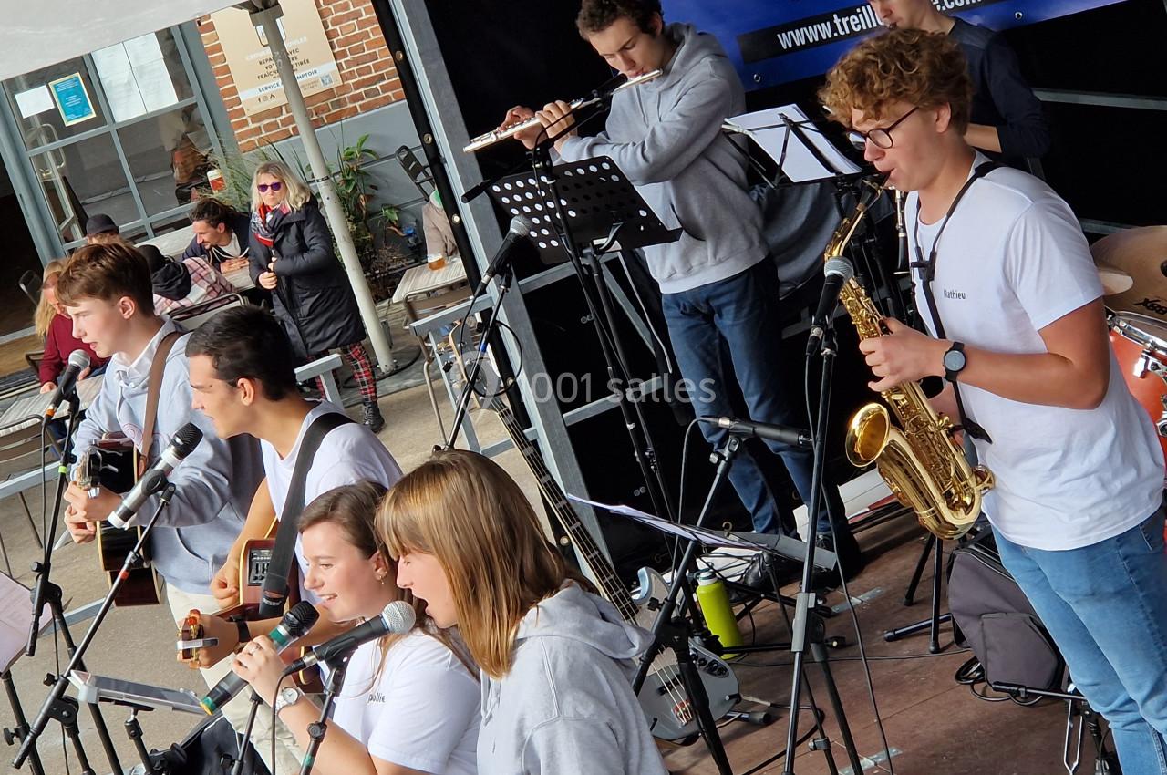 Un groupe de jeunes musiciens joue en plein air avec divers instruments, dont guitares, saxophone et flûte.