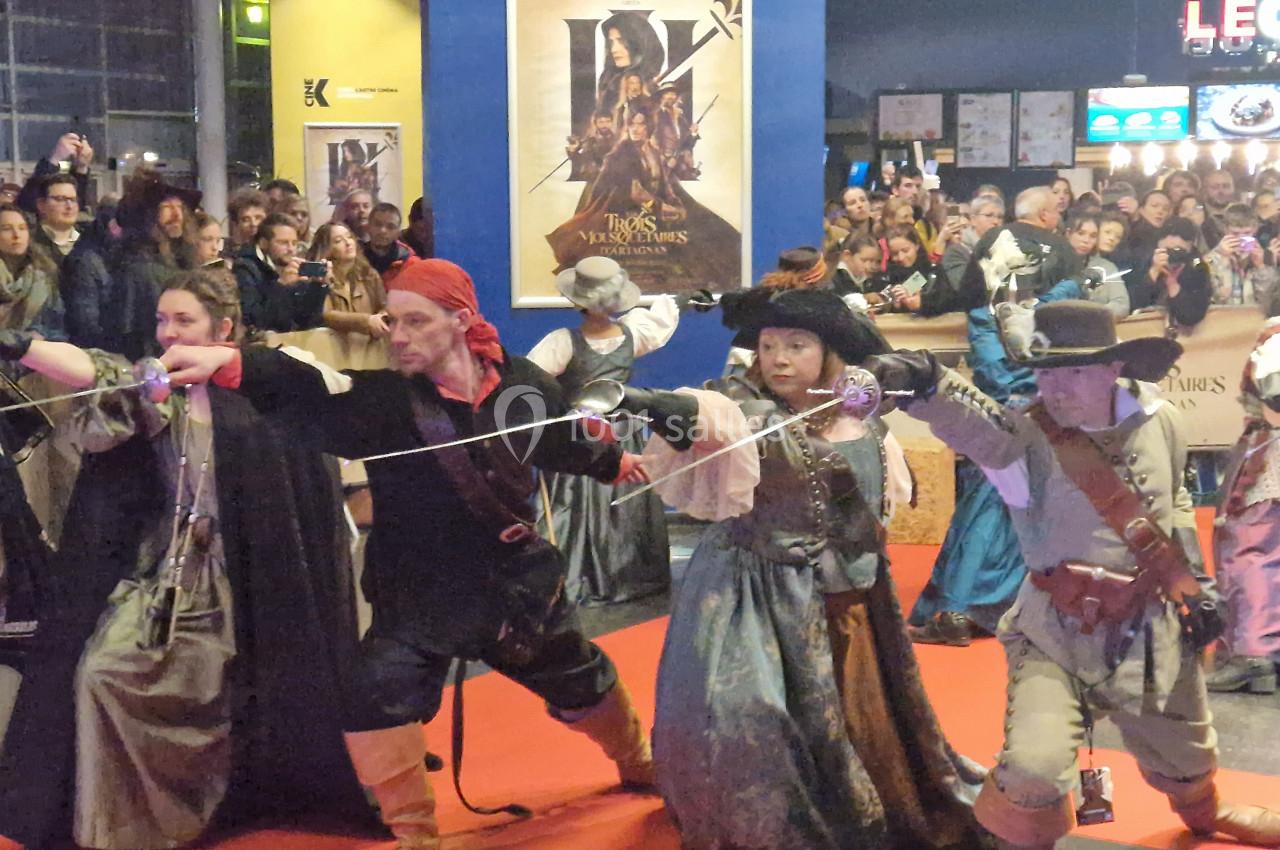 Des personnes en costumes de mousquetaires posent avec des épées sur un tapis rouge, devant un public et des affiches.