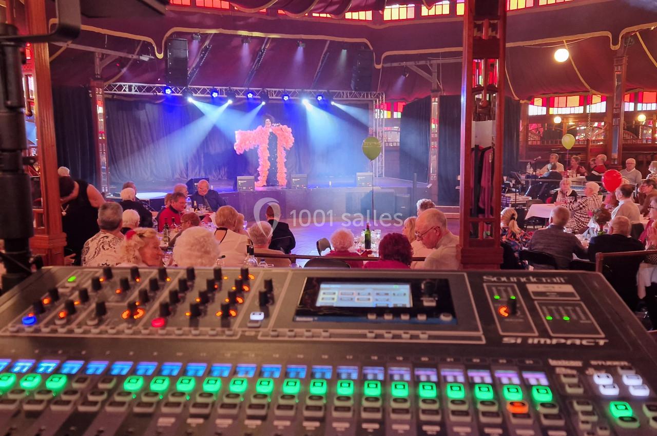 Vue d'une console de mixage audio en premier plan, avec une scène éclairée et un public assis dans une salle de spectacle.