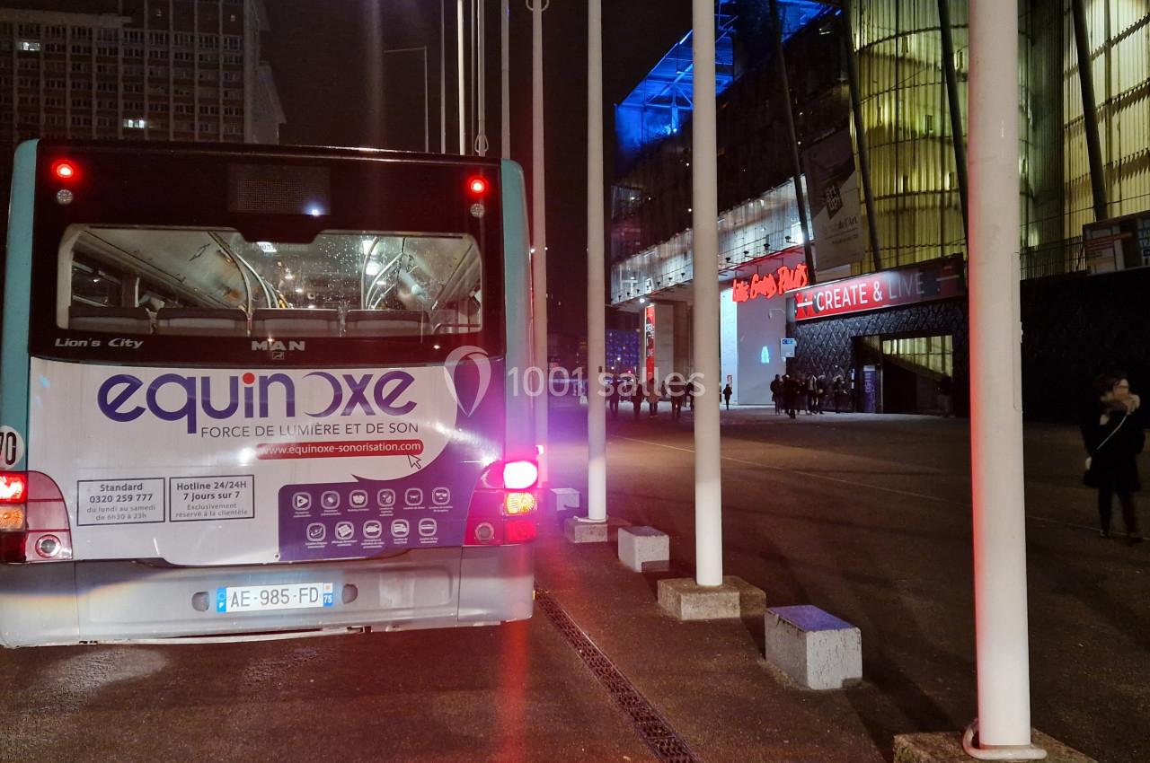 Un bus stationné de nuit devant un bâtiment illuminé, avec des passants en arrière-plan.