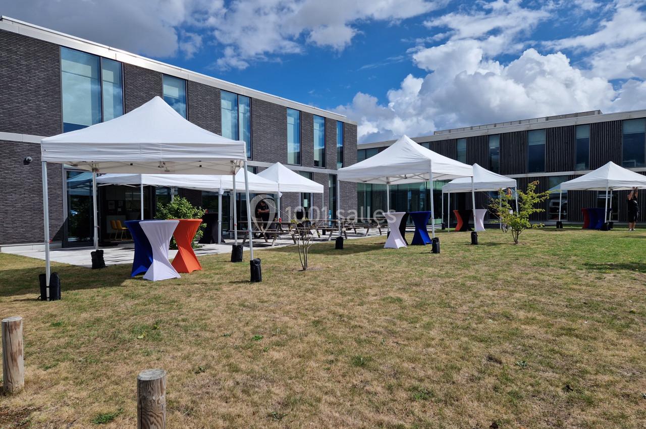 Tentes blanches installées sur une pelouse devant un bâtiment moderne, avec quelques tables et décorations colorées.
