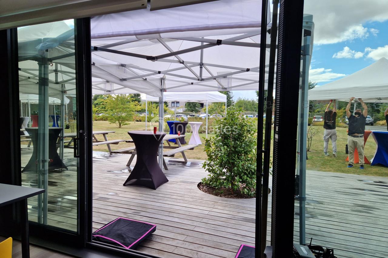 Vue depuis une salle sur une terrasse avec des tables sous des tonnelles blanches et un espace vert en arrière-plan.