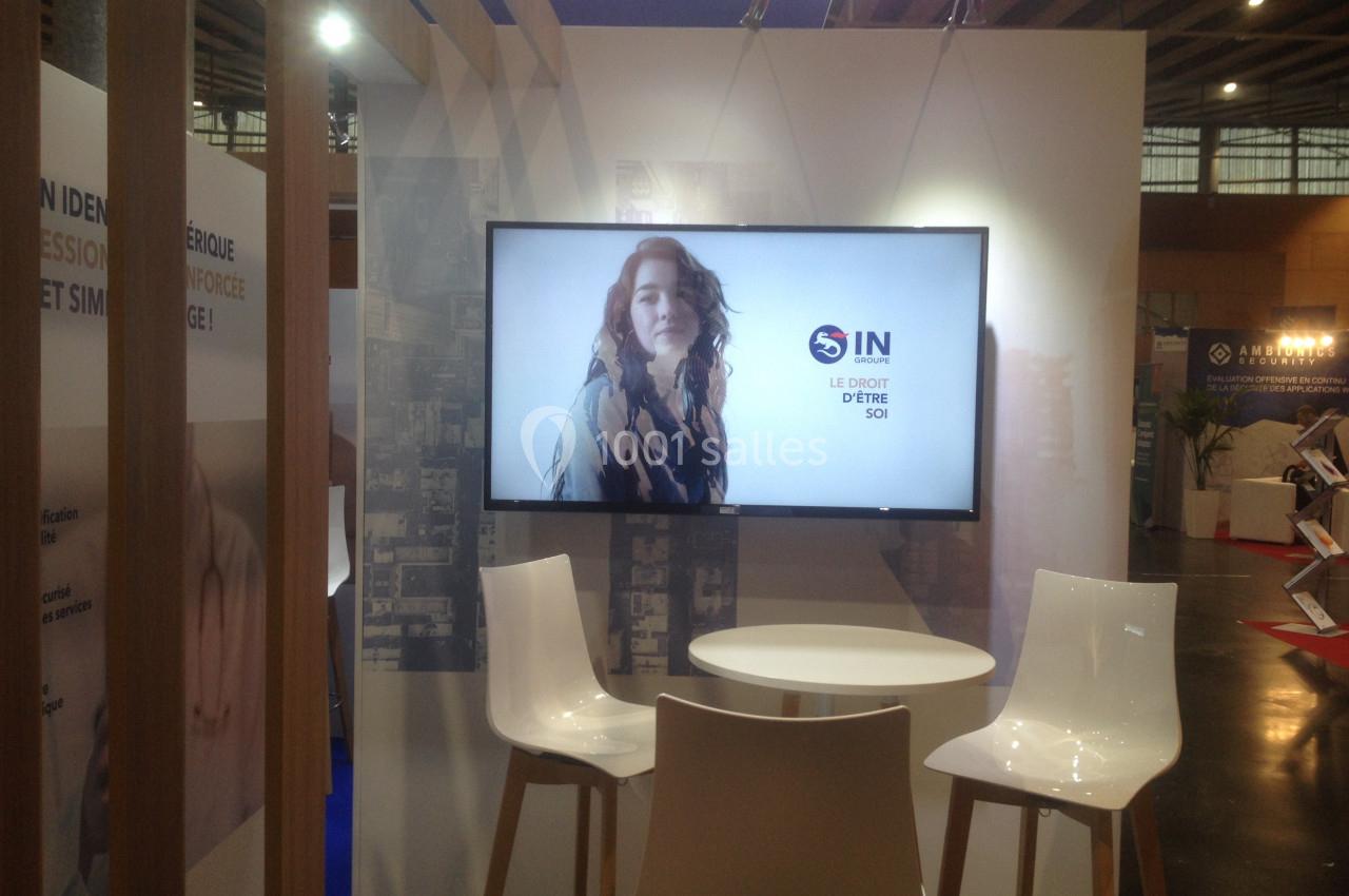 Écran affichant une femme sur fond clair, entouré de deux chaises et une table dans un espace d'exposition.