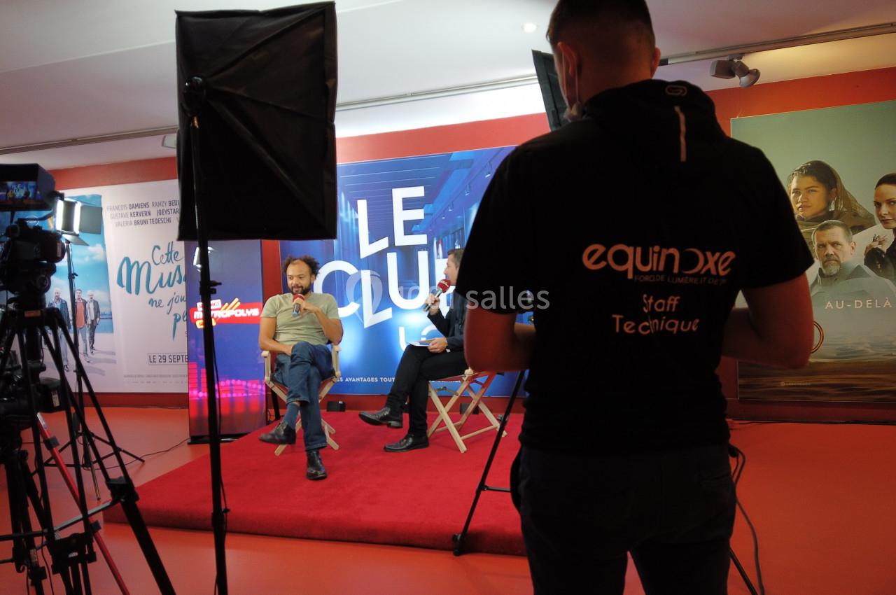 Deux hommes discutent sur un plateau d'interview avec caméras et technicien en arrière-plan.