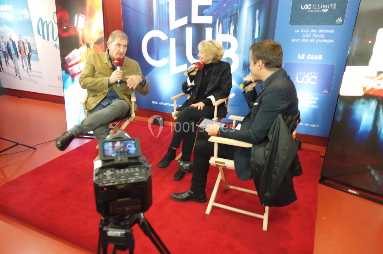 Trois personnes discutent sur une scène avec micros, assises sur des chaises, devant un fond marqué ’Le Club’.