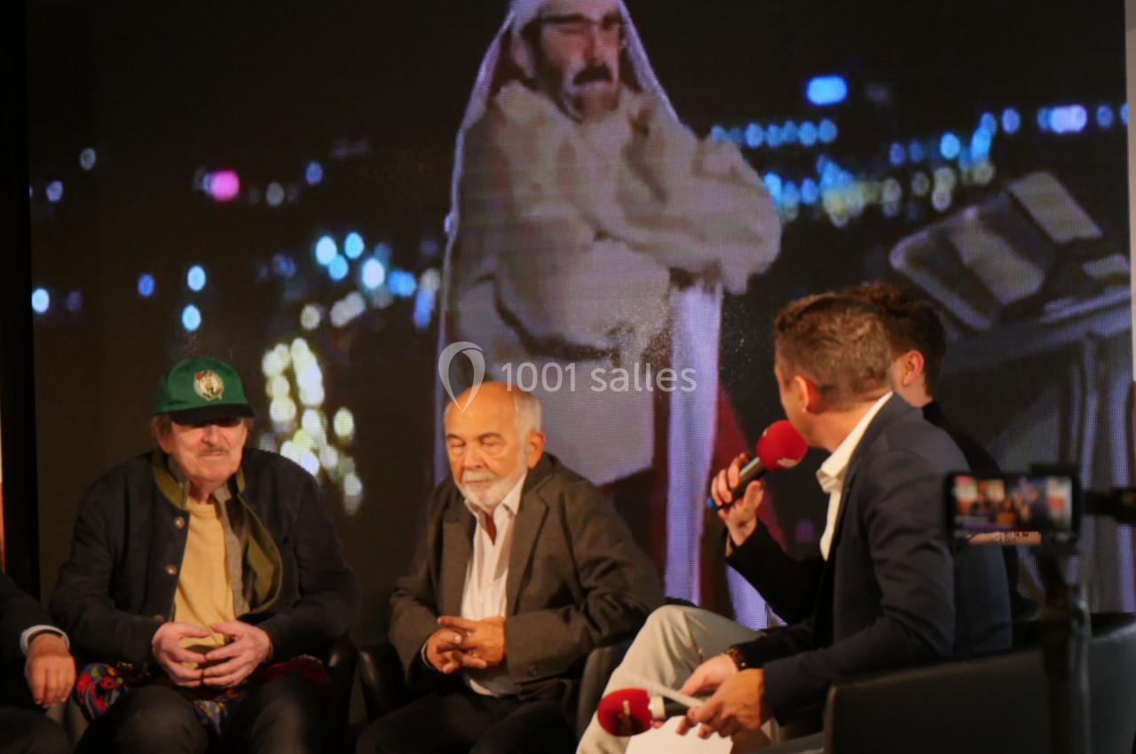 Quatre hommes discutent sur un plateau, avec un écran en arrière-plan affichant une personne en tenue traditionnelle.