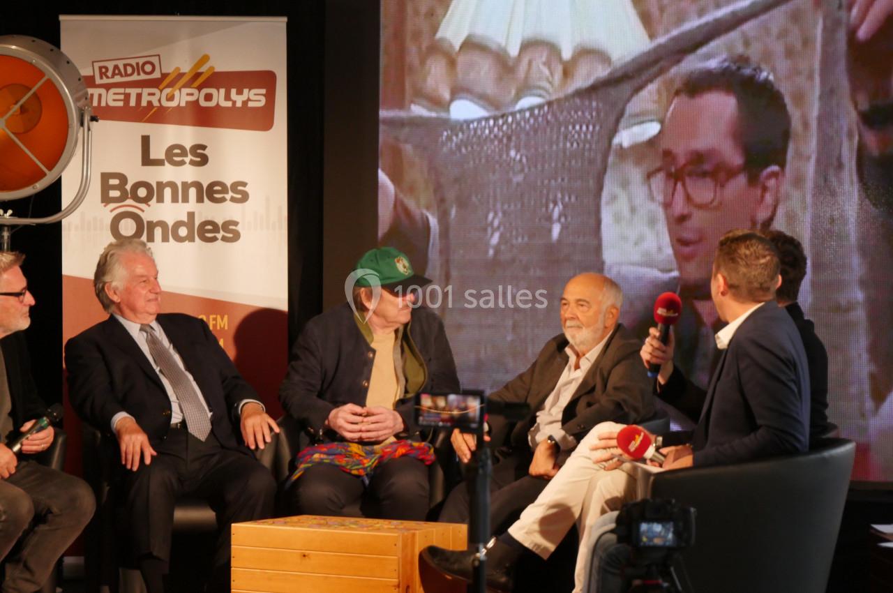 Des personnes discutent sur une scène lors d'une émission radio, avec un écran projetant une image en arrière-plan.