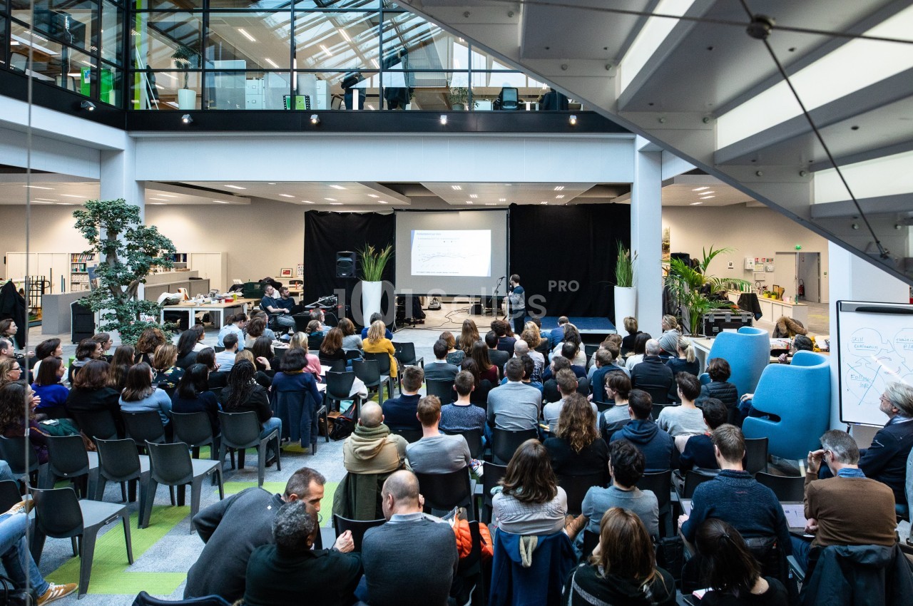 Un conférencier s'adresse à un public assis dans une grande salle moderne avec écran de projection.