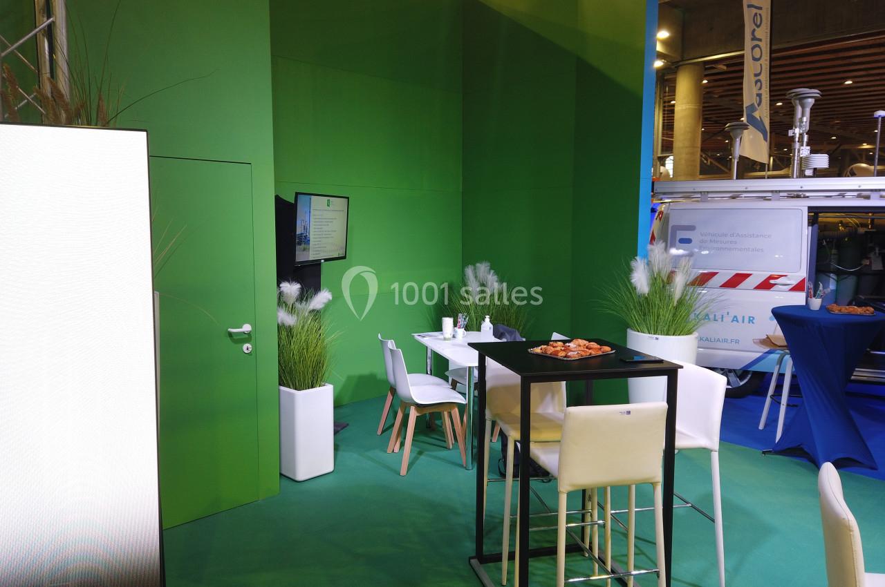 Espace d'exposition avec table haute, chaises blanches, plantes décoratives et écran fixé sur un mur vert.