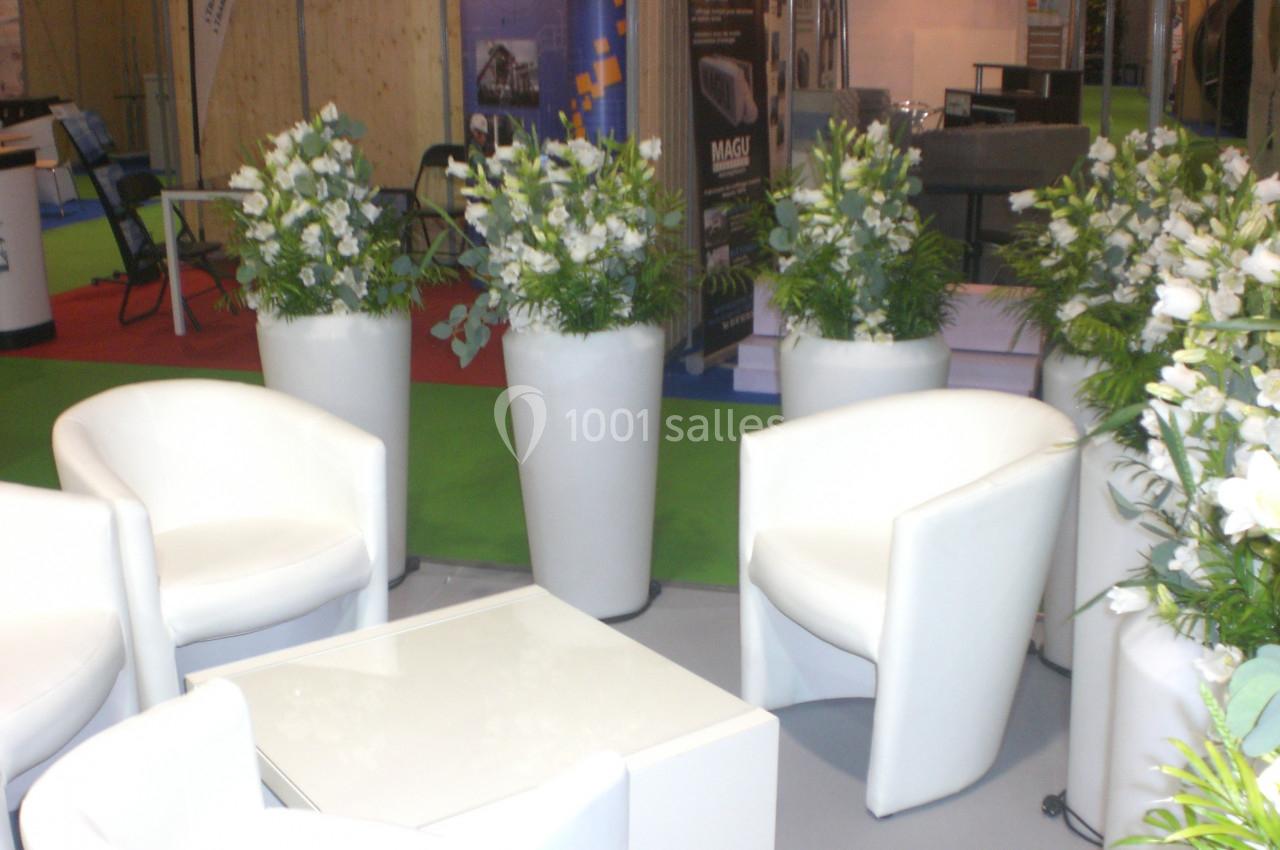 Chaises blanches et tables basses entourées de grands vases avec des arrangements floraux dans un espace d'exposition.