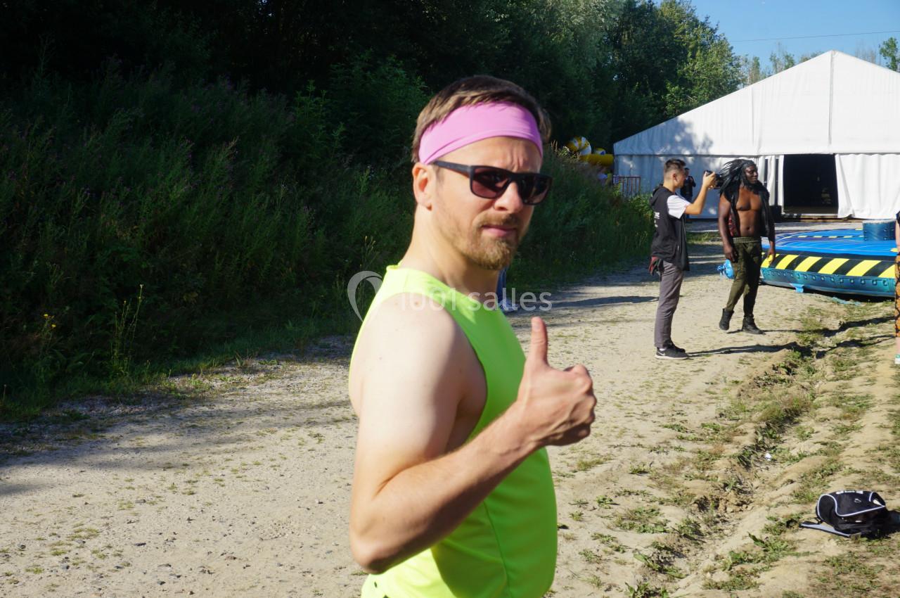 Homme en débardeur jaune et bandeau rose faisant un signe de pouce levé sur un chemin près d'une tente blanche.