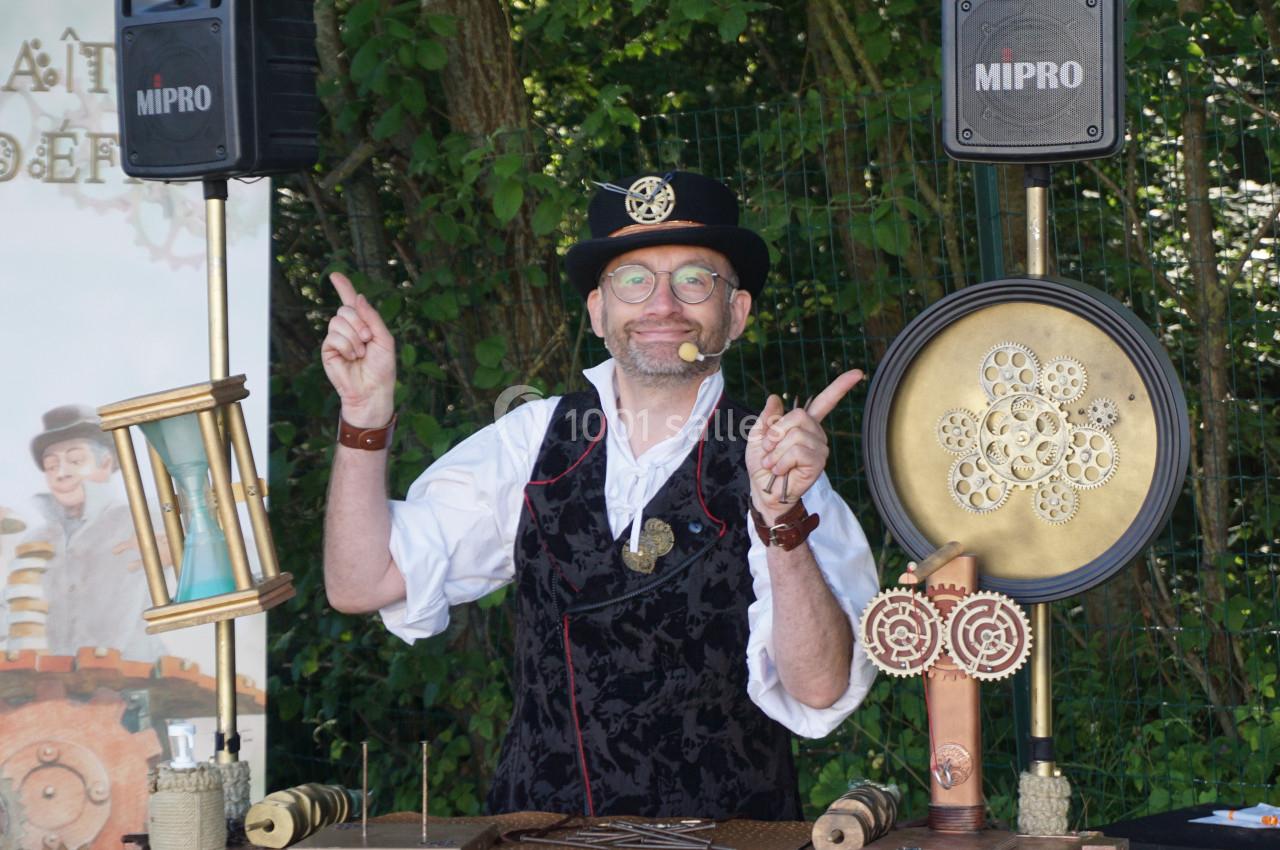 Un homme en tenue steampunk présente un stand avec des objets mécaniques et décoratifs dans un cadre extérieur boisé.