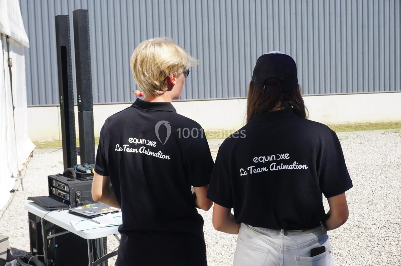 Deux personnes en polos noirs avec l'inscription ’La Team Animation’ de dos, près d'équipements techniques en extérieur.