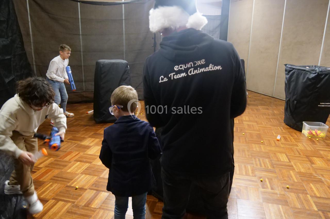 Des enfants jouent avec des pistolets en mousse dans une salle aménagée avec des obstacles et un animateur présent.