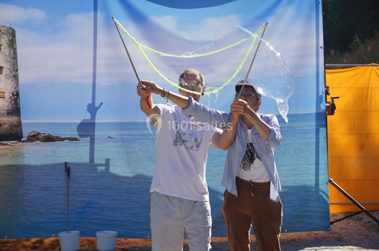 Deux personnes créent une grande bulle de savon à l'aide de baguettes et de cordes, en extérieur près d'une plage.