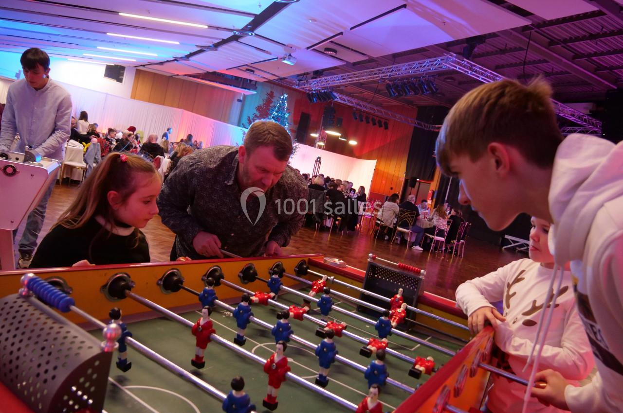 Des personnes jouent au baby-foot dans une salle animée avec des tables et des éclairages colorés en arrière-plan.