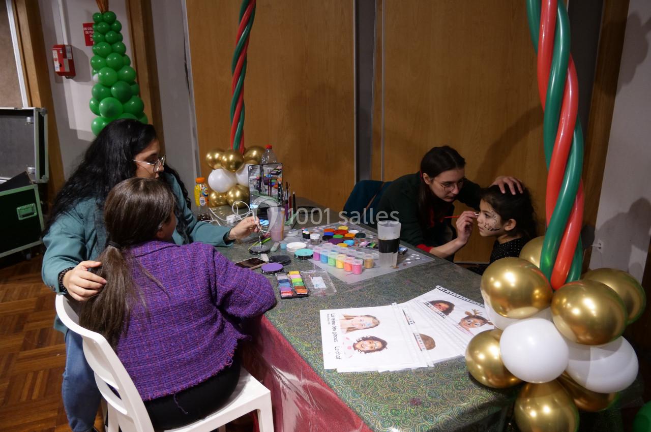 Deux enfants se font maquiller le visage par deux adultes à une table décorée de ballons et de matériel de peinture.