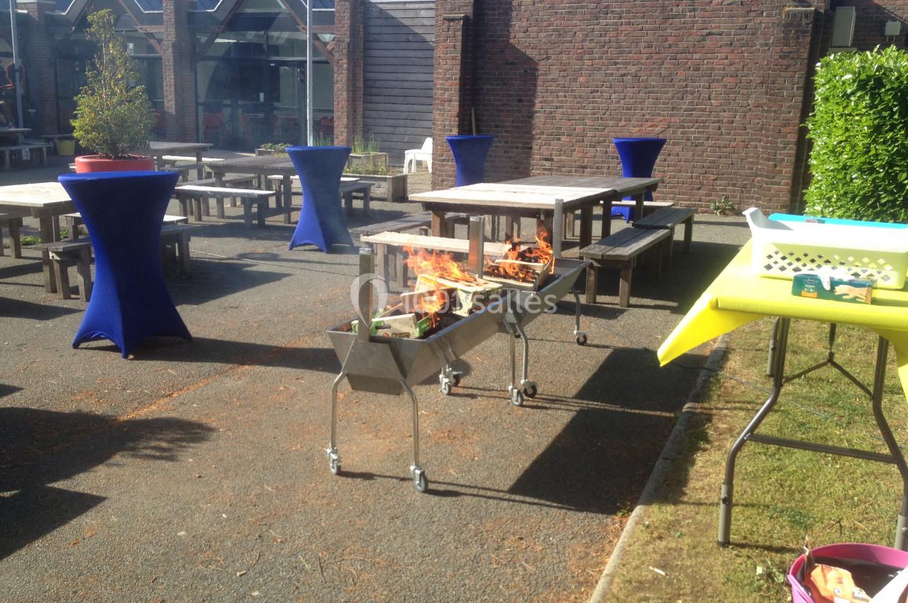 Barbecue en feu sur une terrasse extérieure avec tables en bois et nappes bleues, entouré de bâtiments en briques.
