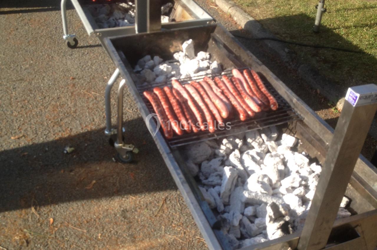 Deux barbecues en métal sur roulettes avec des saucisses grillant sur des braises blanches, en extérieur.