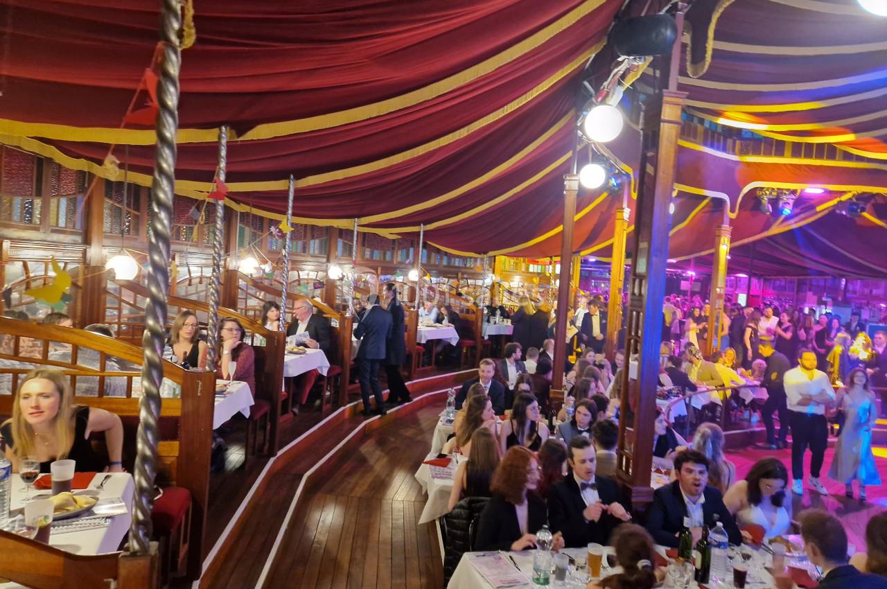 Personnes attablées dans une salle circulaire décorée de tentures rouges et jaunes, ambiance festive et éclairage coloré.