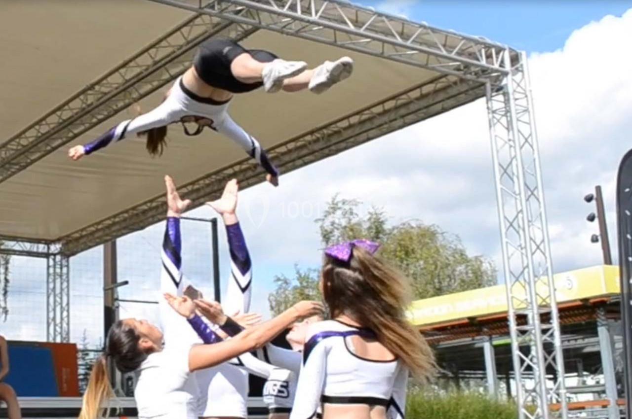 Une équipe de cheerleaders exécute une figure acrobatique avec une voltigeuse lancée en l'air sous une structure extérieure.