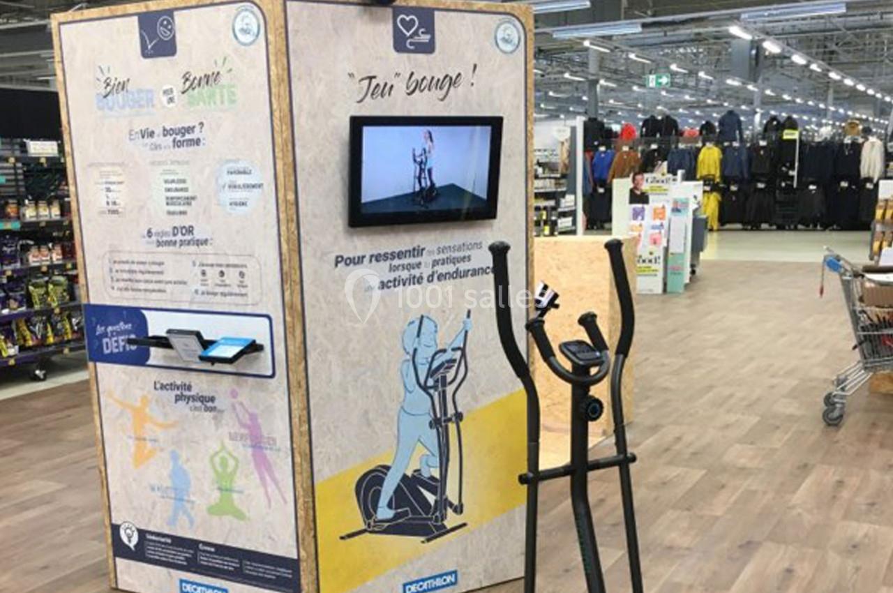 Borne interactive avec écran et vélo elliptique dans un magasin, invitant à tester une activité physique ludique.