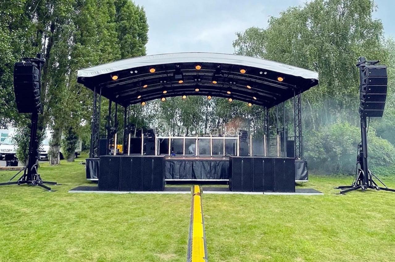 Scène extérieure avec structure métallique, éclairages allumés, installée sur une pelouse entourée d'arbres.